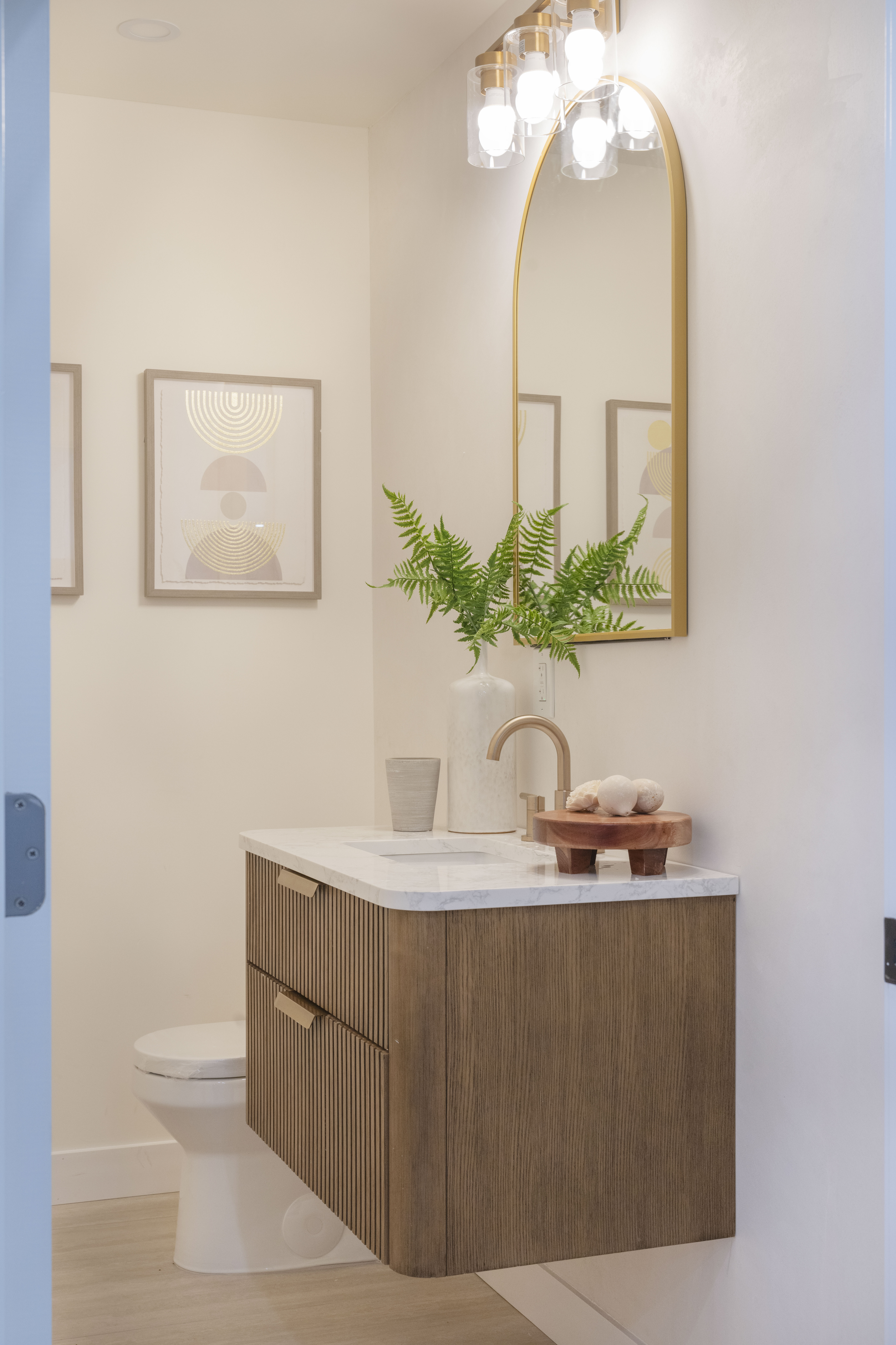 Elegant ADU powder room with gold arched mirror, floating wood vanity, and designer lighting