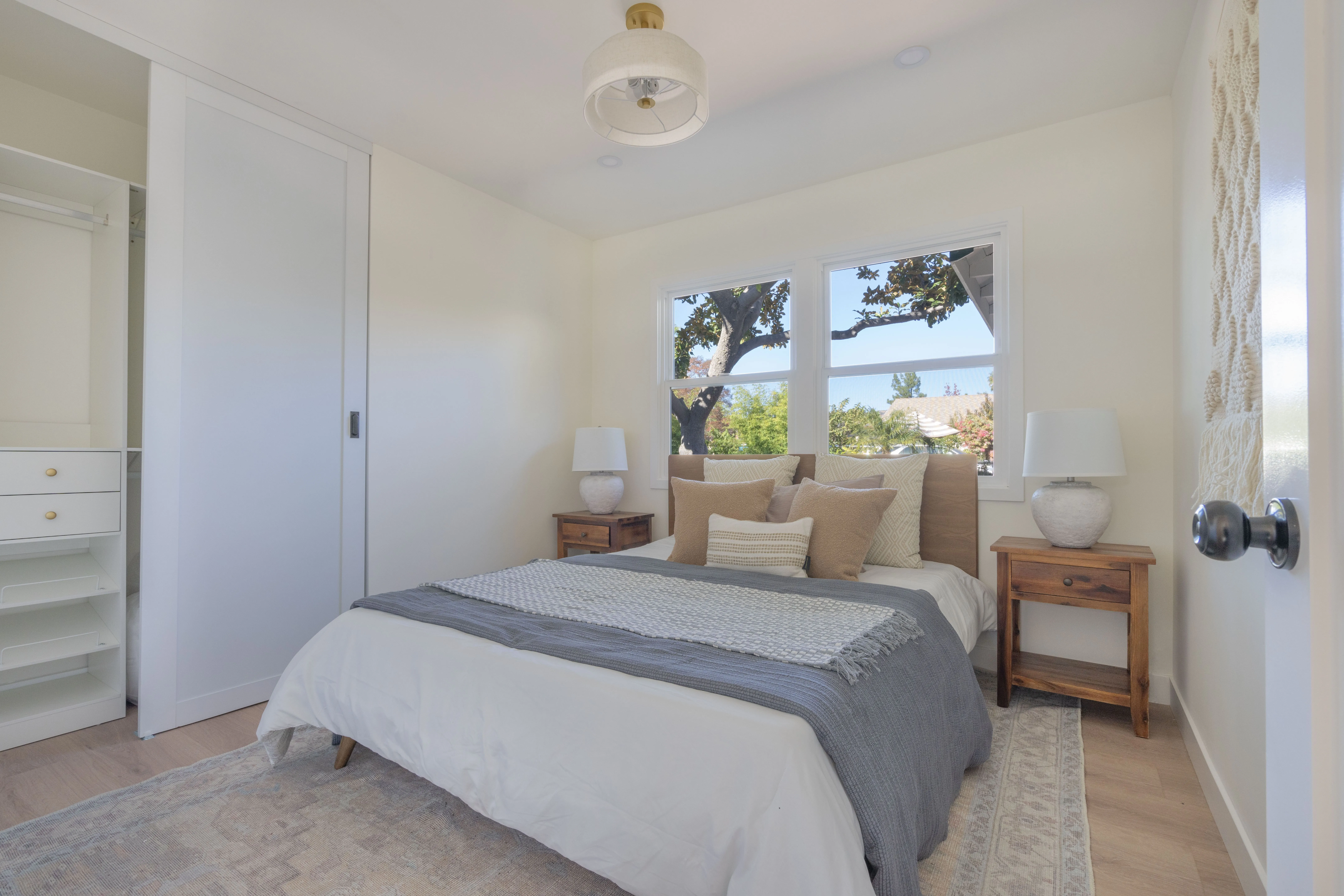 Bright ADU bedroom with built-in closet, natural wood nightstands, and garden view windows